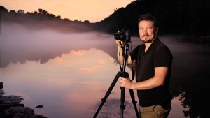 Edward C. Robison III stands in front of a lake at sunset with his camera on a tripod