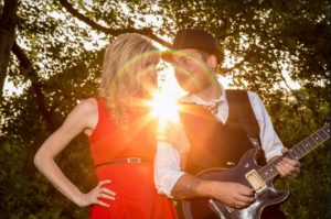 Woman in a red dress next to a man with a guitar. They are standing with the setting sun behind their backs.