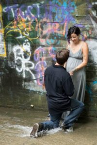 Engagement photo of man proposing to woman. Man is kneeling in moving water while woman stands against a colorful graffiti wall.