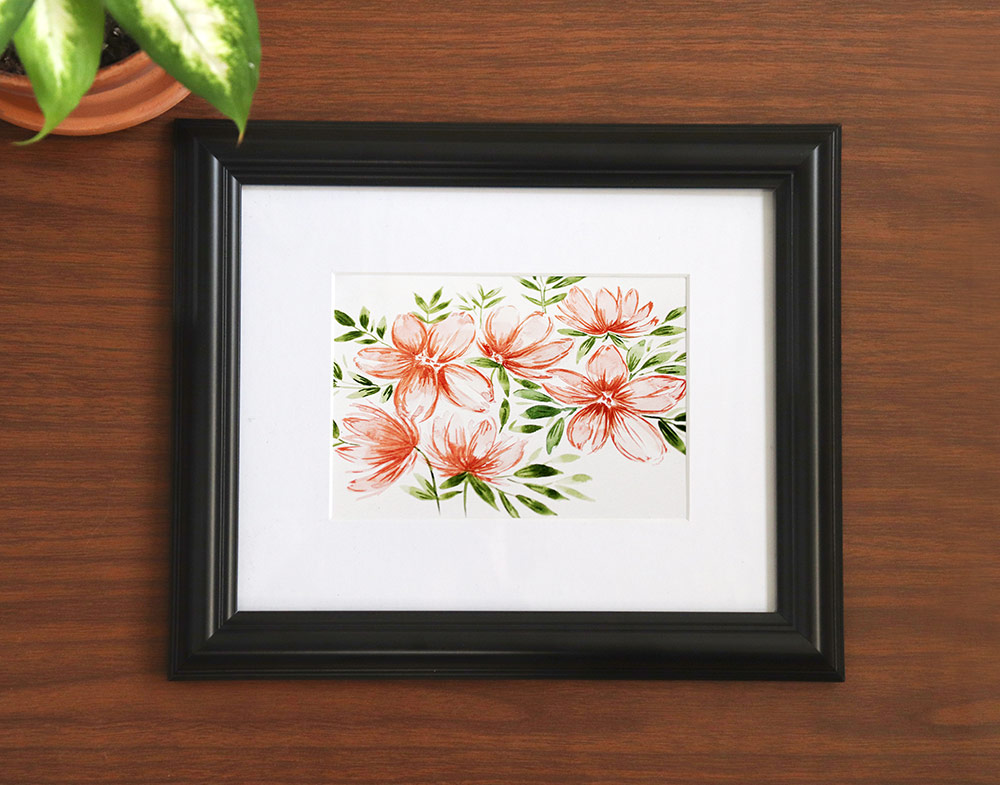 Watercolor floral painting in a black frame with white mat. Sitting on a wooden desk with a plant sitting by the side.