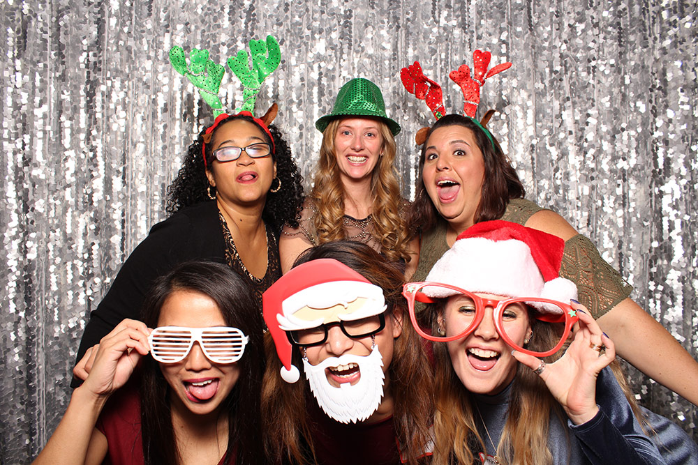 Six people pose in a photo booth in front of a bright, sparkling silver backdrop. They are all wearing funny holiday photo booth props and smiling or making silly faces.