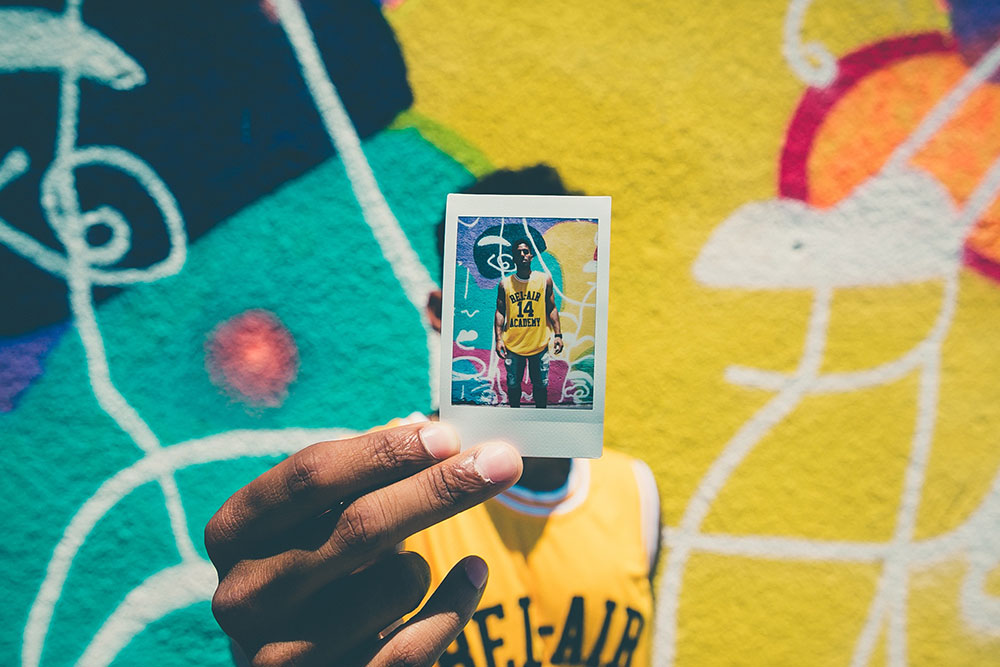 A man holds an Instax Mini picture of himself standing in front of a brightly-colored graffiti wall