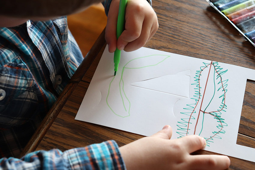 A young child draws on a white paper frame with green and brown markers.