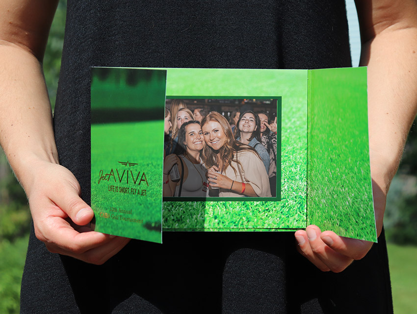 A woman in a sleeveless black dress holds a green golf photo folder on a sunny day in front of a landscaped green yard.