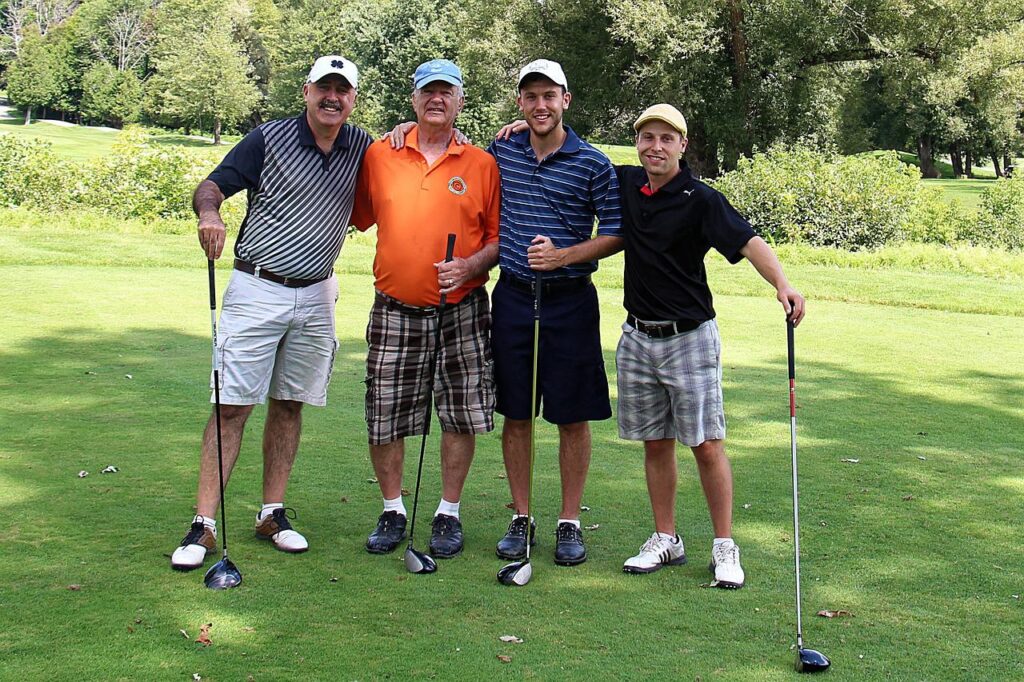 Four male golfers stand together with their golf clubs on the green on a sunny day.