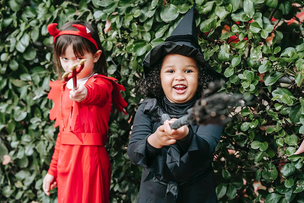 A girl dressed as a devil seriously points her trident at the camera. The other girls is dressed as a witch and is gleefully pointing her broom at the camera.