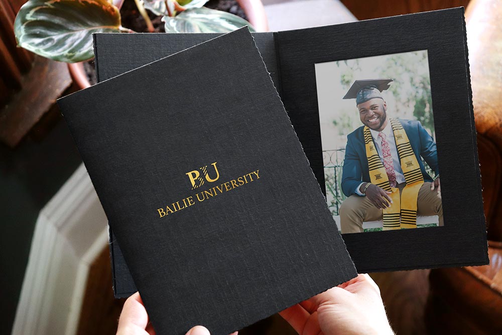 Vertical black portrait folder with ragged edges has a university logo imprinted on the front cover in gold and a university graduate's picture framed inside