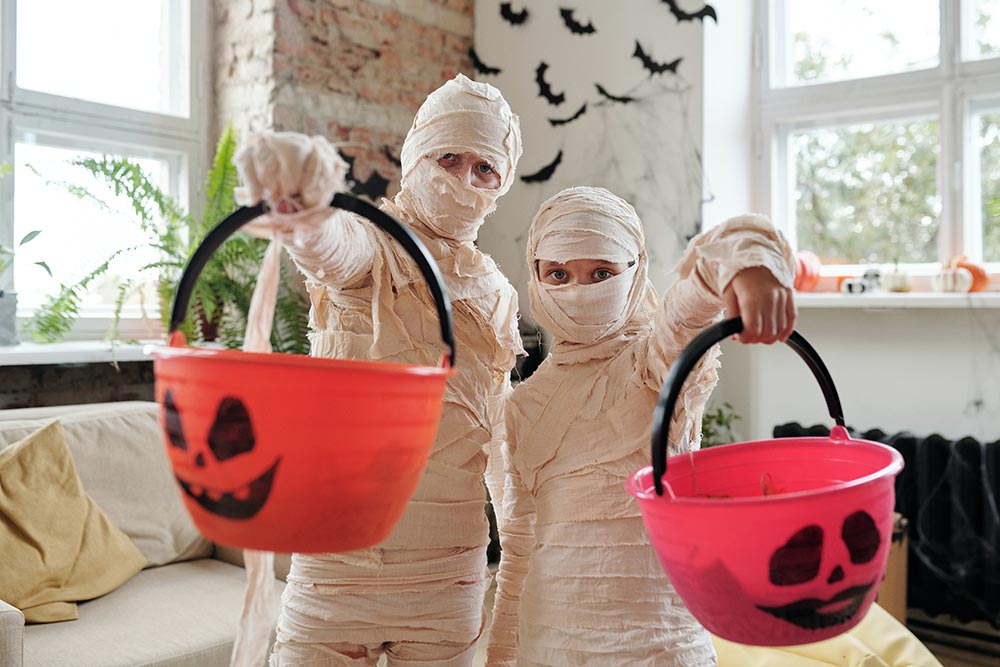 Two kids hold up Halloween pumpkin candy buckets while both are dressed up as mummies