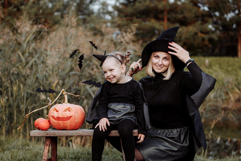 A mother and daughter are outside in witch costumes, sitting next to a smiling carved Jack-O-Lantern for a Halloween photo