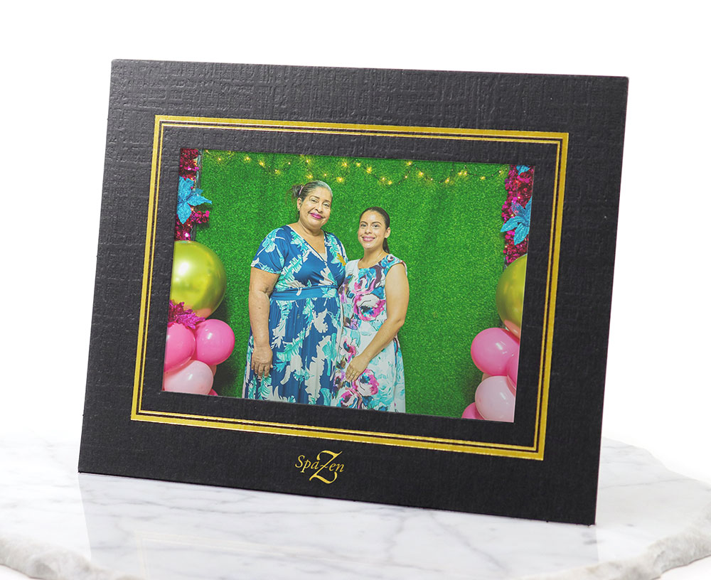 A black paper frame with gold window border accent and logo imprint frames a photo booth print