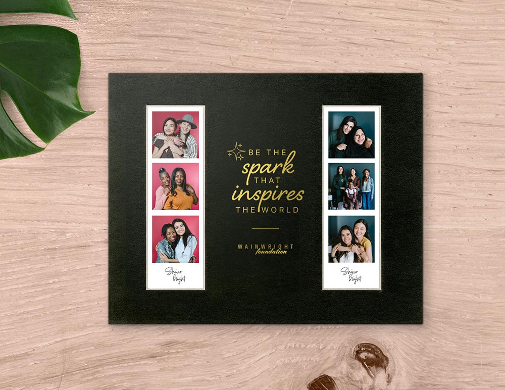 Black picture frame mat holds two photo booth prints. The mat has a gold logo imprint and reads  Be the spark that inspires the world.
