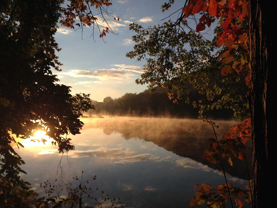 Mist rises off a glass-like lake at sunrise. The landscape scene is framed by a tree on each side in the foreground, with a hill on the side of the lake.