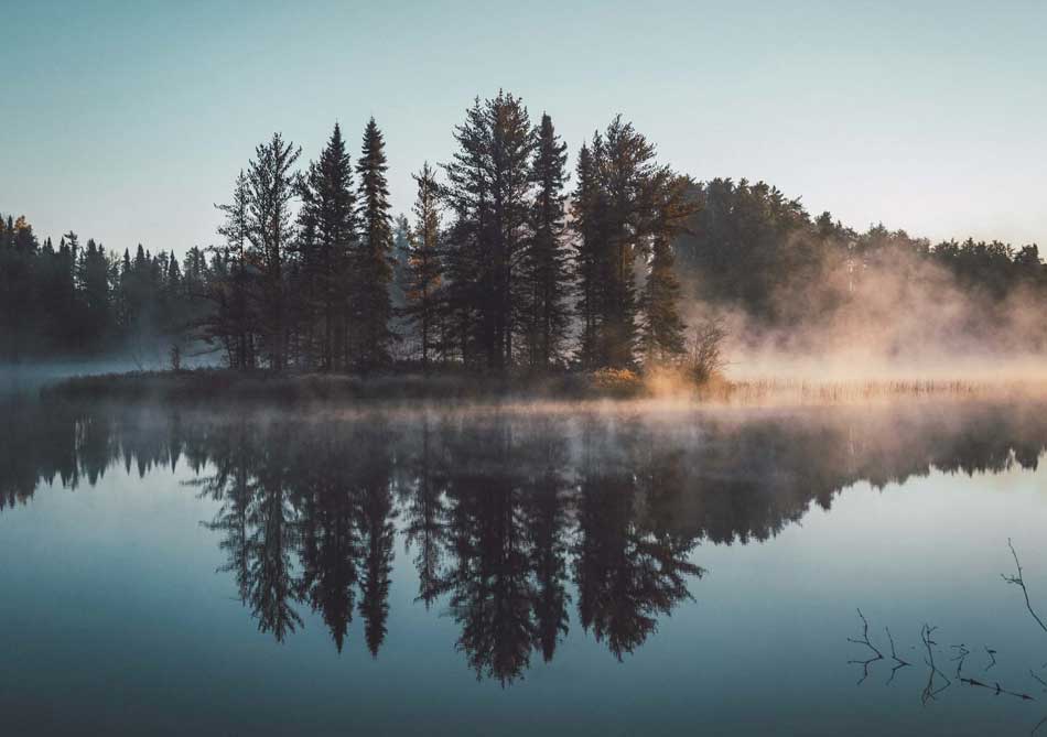 Mist rises off a glass-like lake at sunrise. Evergreen trees on a small bit of land are reflected nearly perfectly in the water.