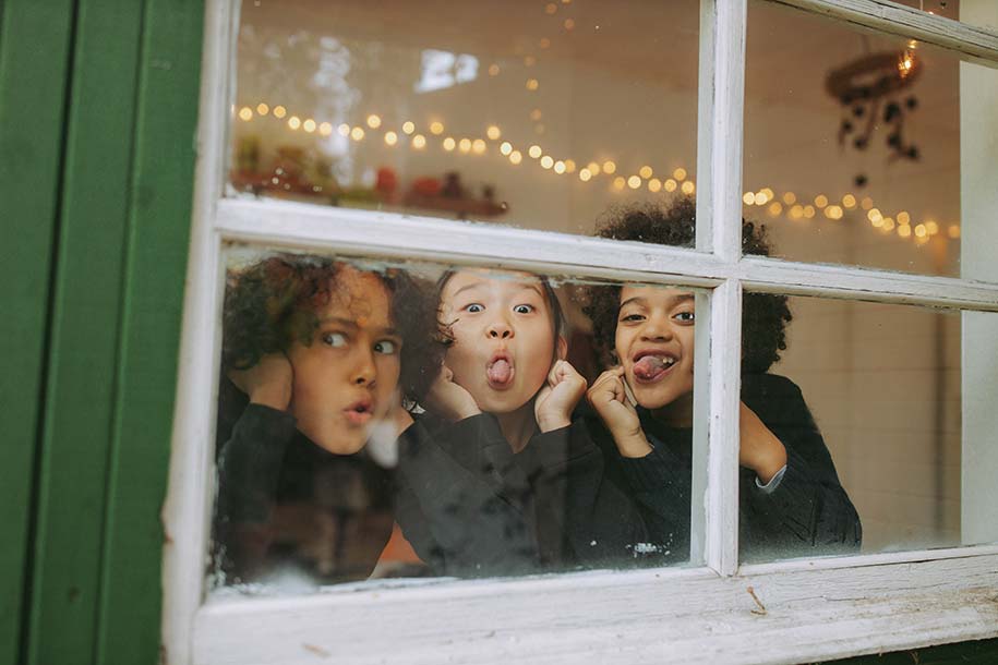 Three young children are inside a house, making silly faces out the window at the photographer.