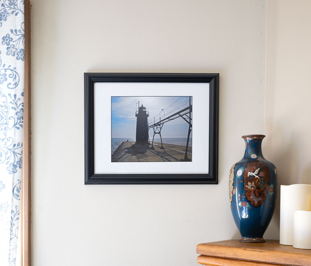 Black frame with white mat board holds a photo of a sun backlit lighthouse and pier. The frame hangs on a wall between a window with a light curtain and a fireplace mantle that's holding a decorative vase and some candles.