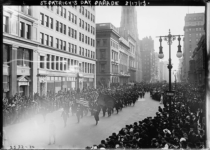 Antique black and white photo of a Saint Patrick's Day parade in a city, on a crowded street. Tall buildings line each side.