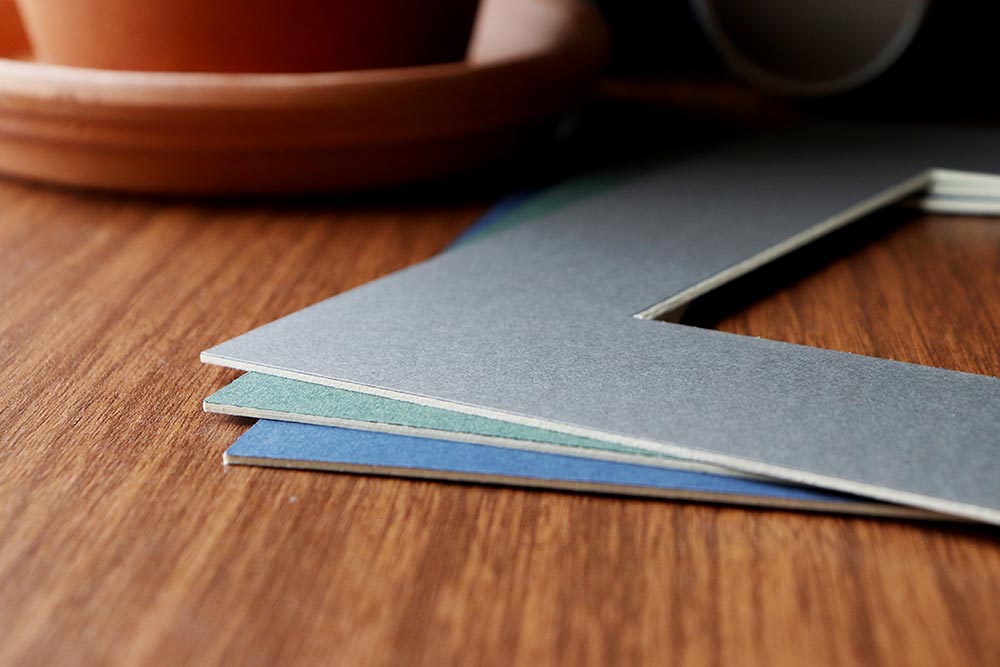 Blue, green, and gray mat boards lay on a wooden desk with a clay flower pot blurred in the background. The image shows the thickness of the mats.