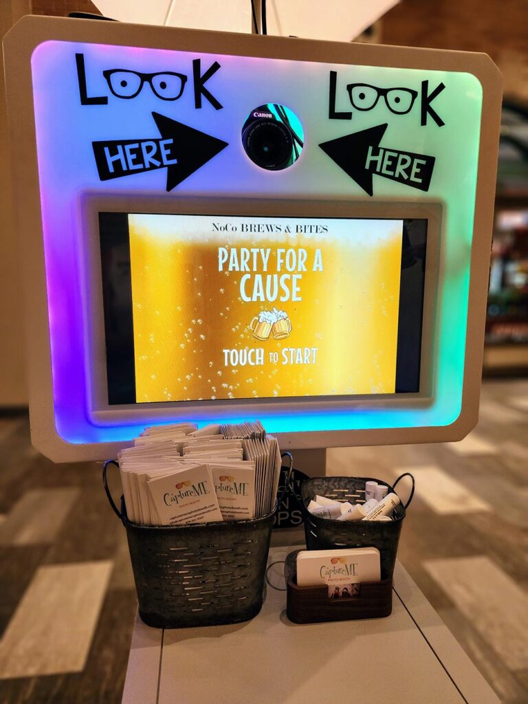 A dSLR photo booth set up with a touch screen below the camera hole. Photo booth folders, lip balm, and business cards sit on a table beneath the touch screen.