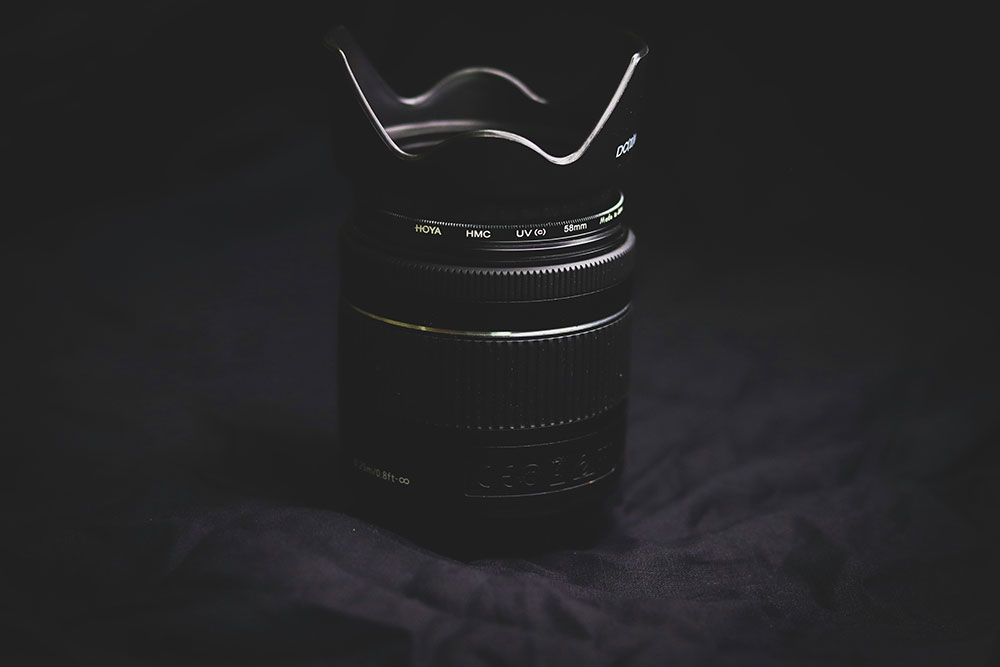 Hood is attached to a camera lens, which is sitting on a dark blanket with subtle light hitting the lens.