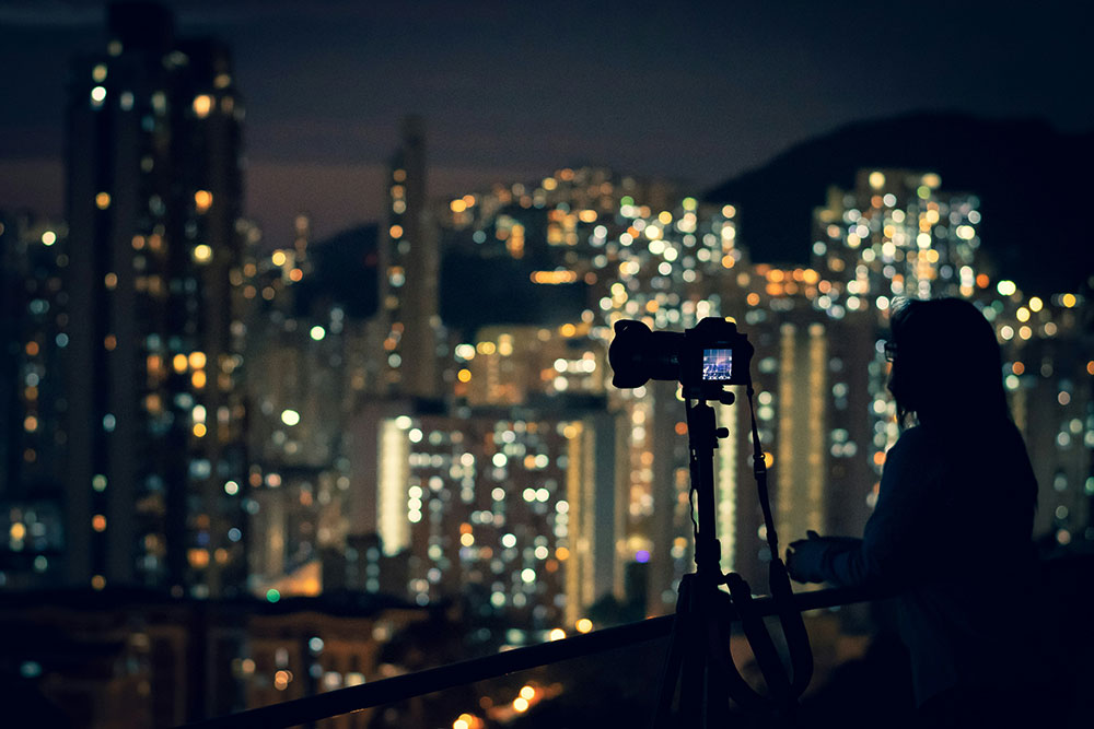 A photographer stands next to their camera on a tripod, which is pointed at skyscrapers at night.