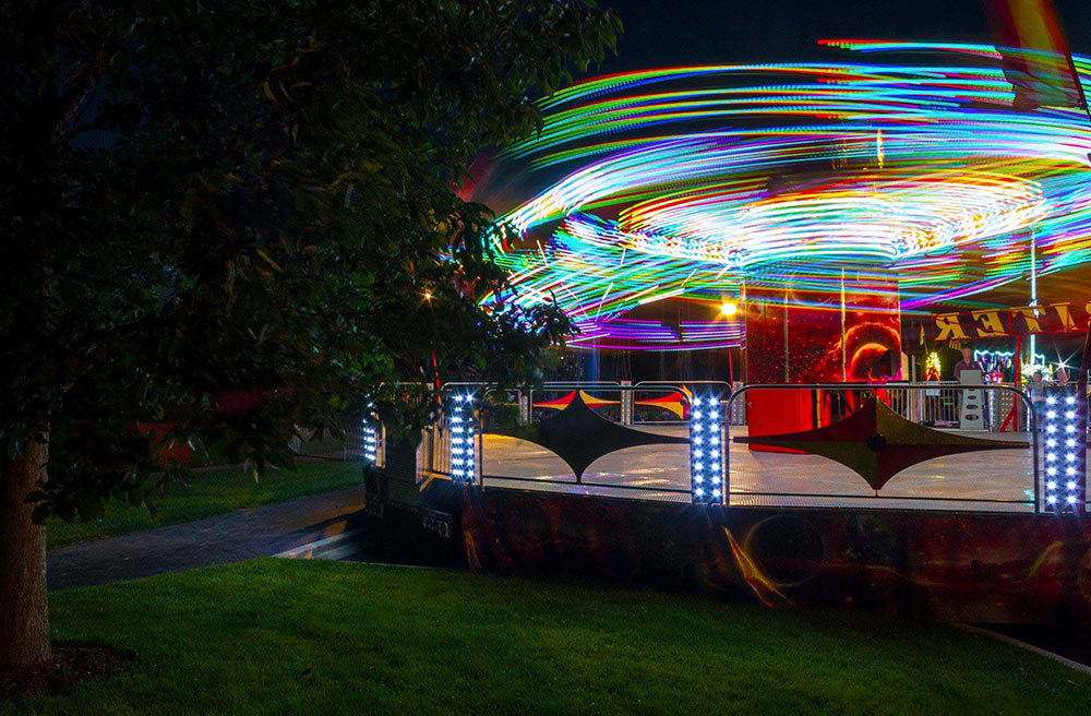 A lit-up carnival ride spins at night in a park, revealing colorful trails of light against the dark night sky.