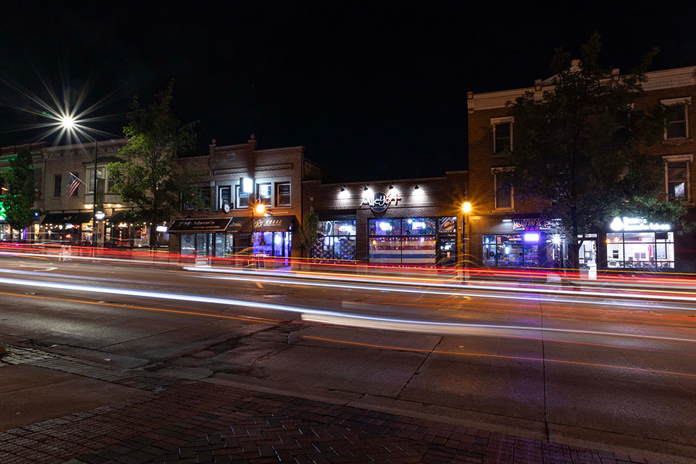 Car headlights and taillights streak down a city street at night, with lights from the shops illuminating the area.