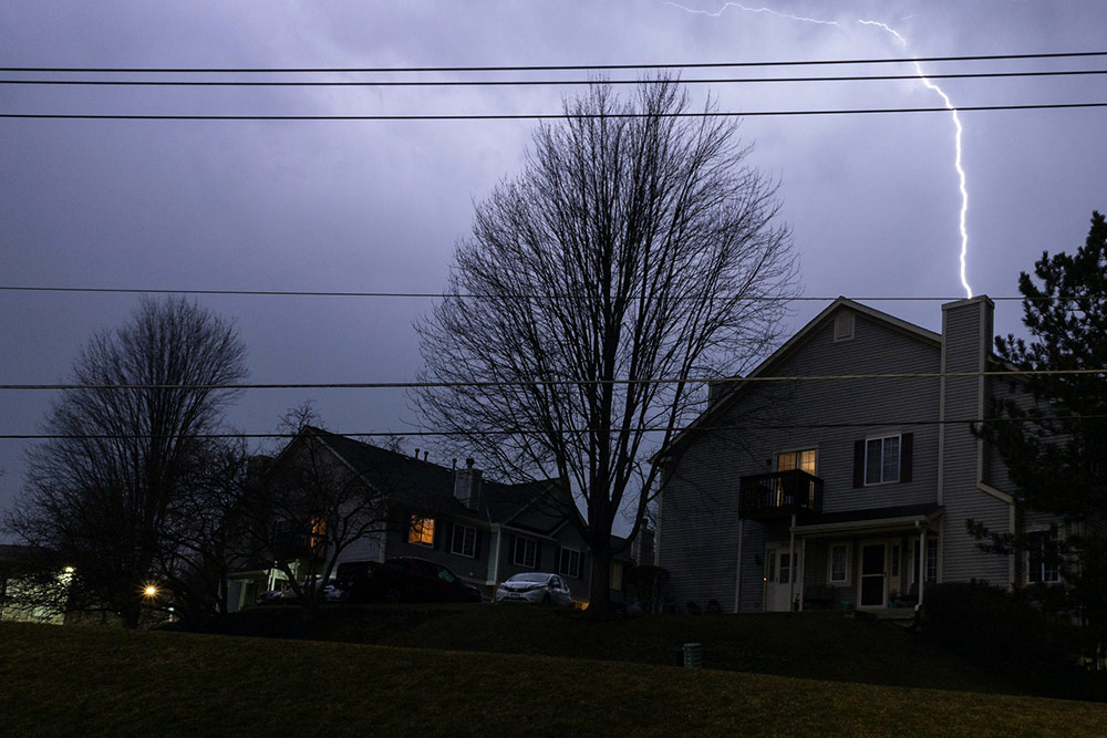 A bolt of lightning flashes behind houses and cars.