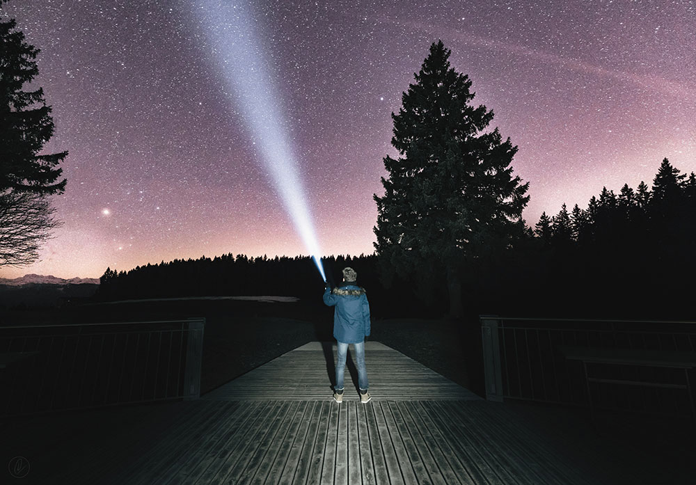 A man stands on a wood pier, pointing a bright flashlight at the star-filled night sky. Bright lights illuminate the man from behind.