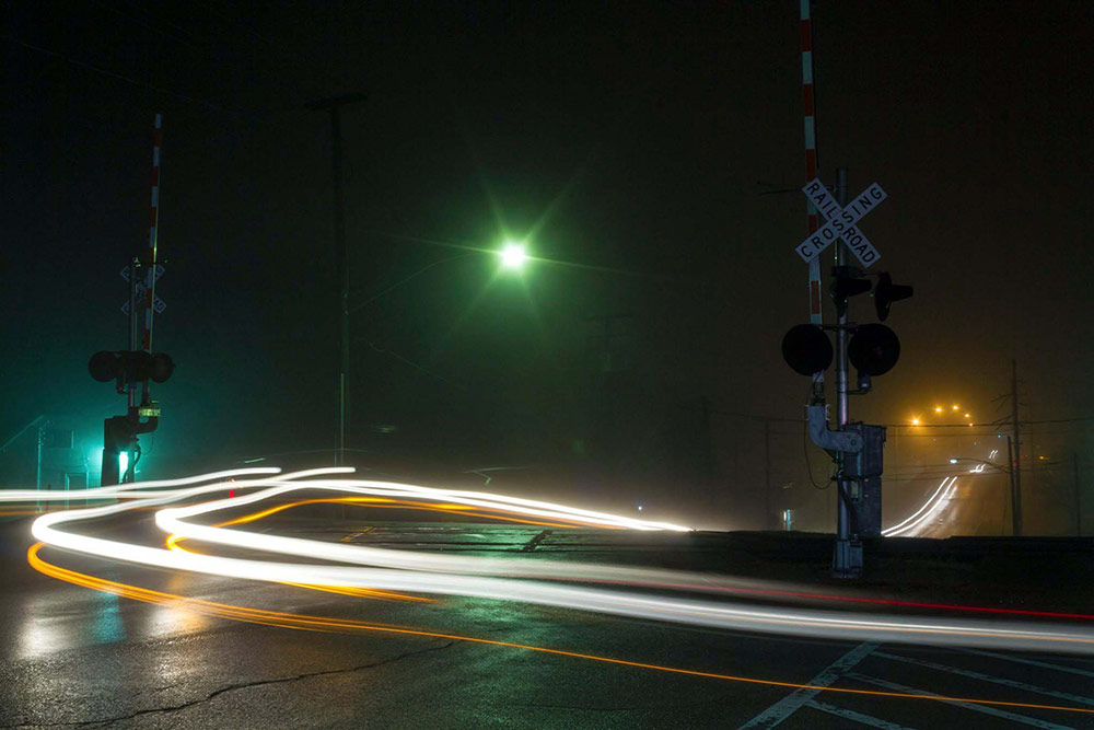 Car headlights trail in a long-exposure photo taken on a misty night at a railroad crossing.