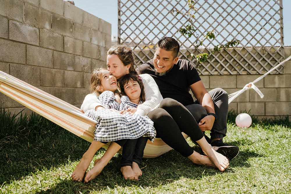 A mom, dad, and their two daughters sit in a hammock in their backyard in the bright sunlight. It is a casual family photo.