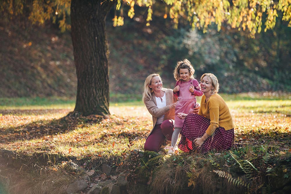 Two women and a young girl smile as the late afternoon sunlight hits them from behind. They are sitting in the grass, under a large tree.