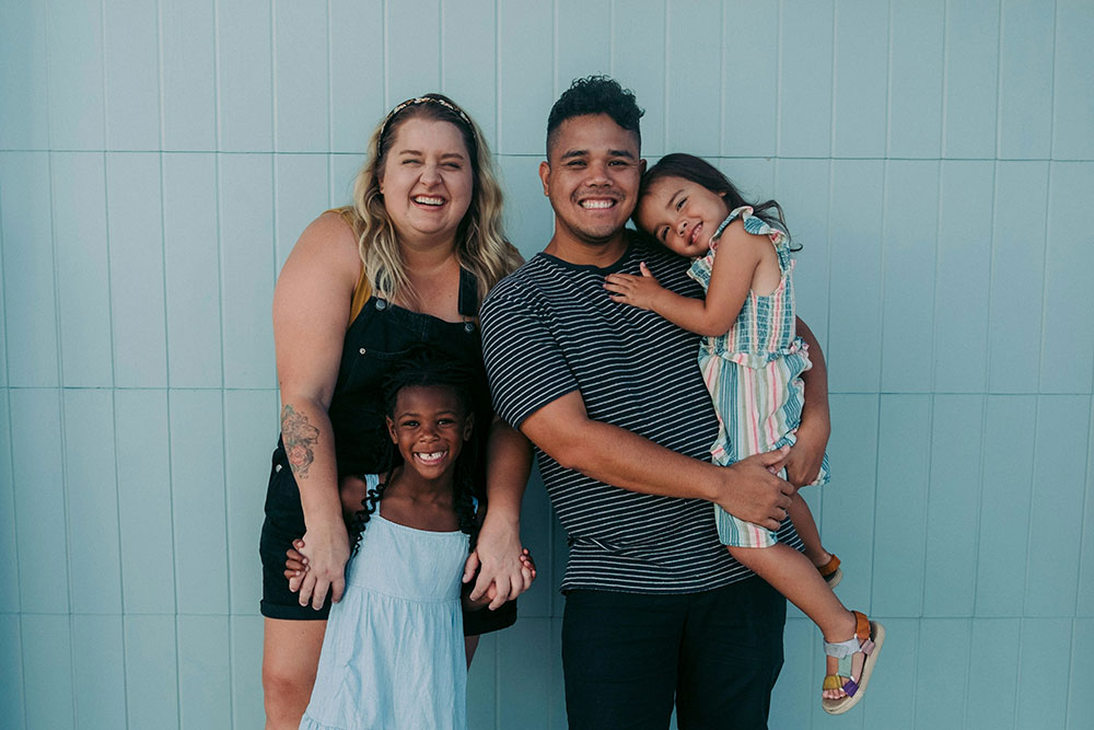 A mom, dad, and their two young daughters smile as they stand against a light blue wall.