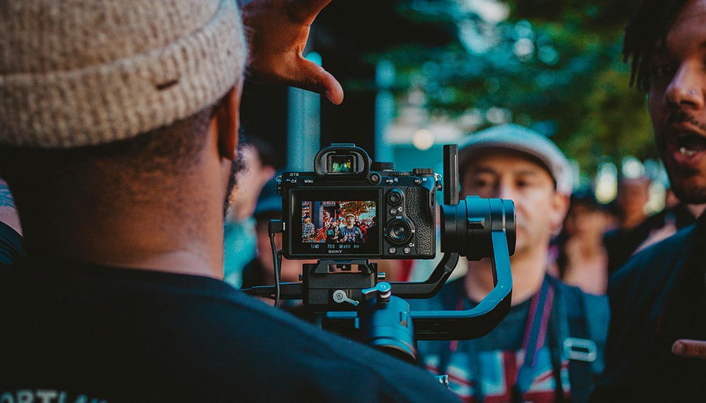 A videographer looks at the camera screen to get the shot of a man standing on a crowded sidewalk.